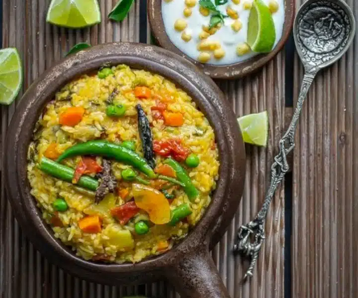 A shot of a traditional Rath Yatra food Khichdi served in a round bowl on a wooden holder. There's an antique spoon to go alongside the vegetables assorted vibrant Khichdi. There are lemon's and leaves and a raita or dahi like substance in a smaller bowl and all these adorn the stripe textured wooden table in frame.