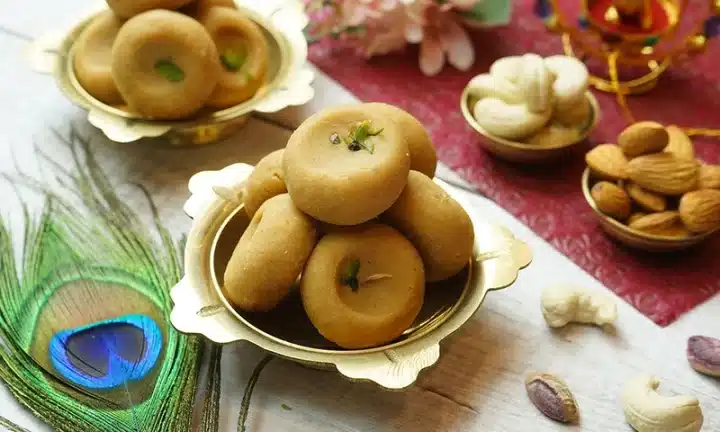 A shot of a traditional Rath Yatra Chappan Bhog food Mathura Peda stacked on top of each other served on a mini round plate. The dish is dummied by another dish on the side. There are also a Peacock's feather, Almond, flower and Cashew in frame.