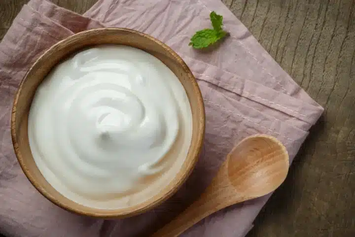 A shot of a traditional Rath Yatra Food Mitha Dahi in an appealing minimalistic serving along with a spoon, a small leaf and textured wall. The brown bowl that the Mitha Dahi is served on is sitting on a pink-ish cloth.