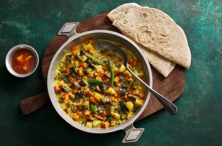 A shot of a traditional Rath Yatra Food Santula with an appealing vibrant look of the vegetables served in a round bowl alongside Chapatis or Rotis. The bowl and the Chapatis are served on a brown wooden holder with a red liquid substance on the side.