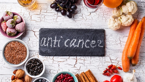 An Image that has a black slate in the middle that has "anti cancer" written on them. It is surrounded by different vegetables, fruits and herbs.