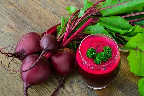 An image of raw beetroot laying on the side of a glass of Beetroot juice on wooden textured background.