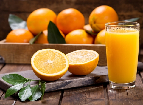 An image of Stanol or sterol-fortified orange juice. Juice is served in glass and fresh and ripe orange are cut in half on the side on chopper. There's also a small leaf branch on the chopper and there are many more bright saffron out-focused or blurred oranges on the back in a wooden container.