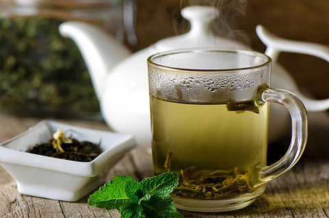 An image of green tea in a cup with kettle. There's also raw tea and a leaf in the image.