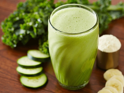 An image of a green smoothie served i a transparent glass with interesting pattern on glass. It is surrounded by different green or healthy veget
