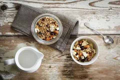 An image displaying fortified cereals from the top angle in one bowl and jar. There's also a spoon, a cloth and a liquid jar on the wooden bench.