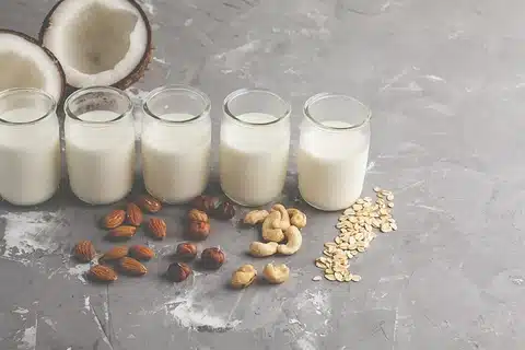 An image of 5 glasses of different types of milks surrounded by the key-elements they are made of such as almonds.