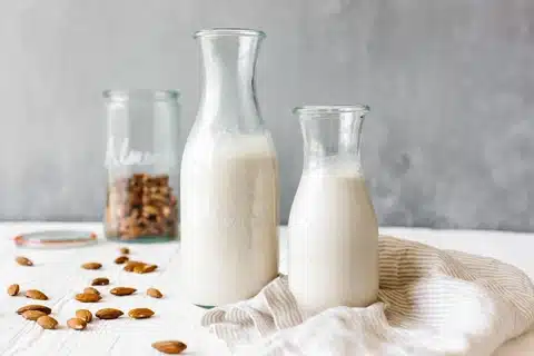 Image of two milk bottles in center. There's a piece of cloth on their right side while on their left is a trail of almonds that leads to the almond jar in the back.