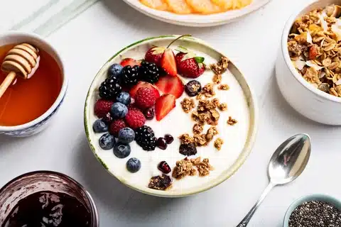 Mouth Watering Yogurt served in a bowl mixed with various fruits like strawberries, grapes and a lot more. There are other bowls bleeding out of the image that contain the items served on top of Yogurt.