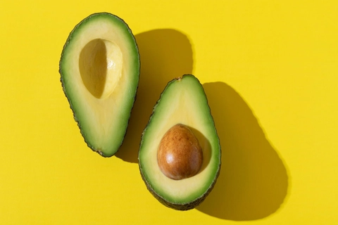 An image of Avocado cut in half, one piece doesn't have the seed while the other has the whole seed on it. Avocado pieces lay on a yellow background with their shadows on their right side.