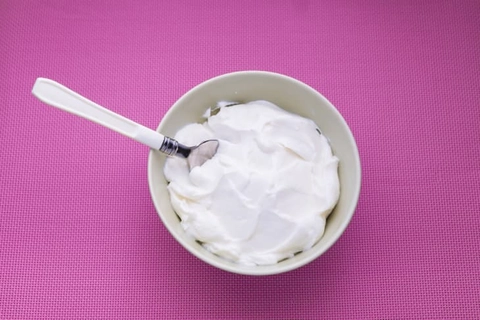An image of a slightly pink-ish yogurt in a bowl with a spoon in front of a violet or bright purple background.