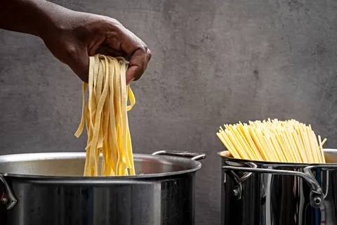 Image of dried pasta in a pot and another pot with long cylindrical noodle like stuff in hand being dropped in or picked out of second pot.